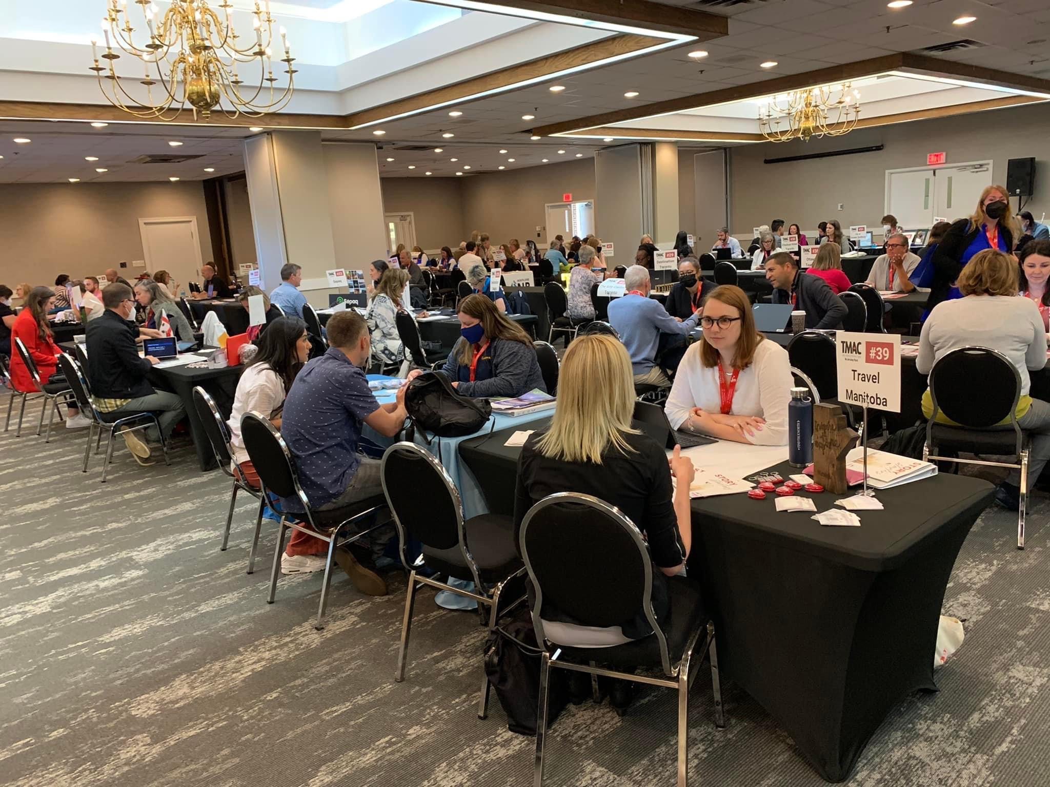 Large room with people sitting across from each other at tables for the TMAC Media Marketplace.