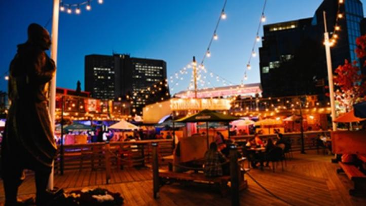 String lights over tables set up on a patio at night with tall buildings in the background.