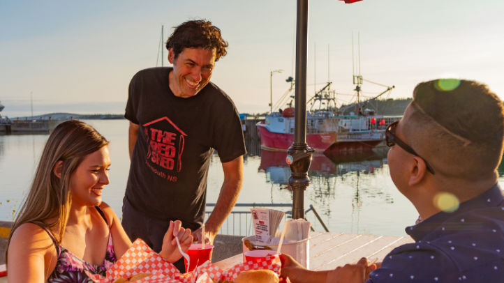 Two people dining outside on a picnic table by the wharf while a server checks on them