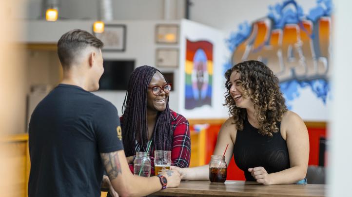 Three people sitting at a table smiling with drinks in mason jars. 