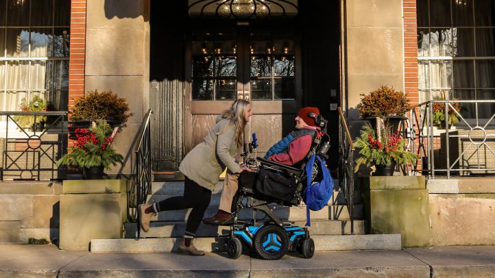 Two people, one of them in a wheelchair, in front of a building outside looking at each other